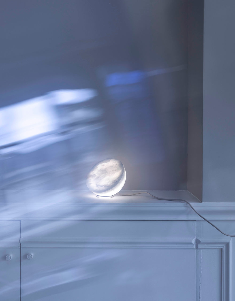 MOON Sixteen Alabaster lit on a white cabinet, daylight across the wall